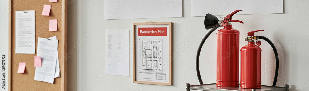 Obraz premium Emergency Preparedness: Two red fire extinguishers stand ready, mounted near a corkboard with notes and a framed fire escape plan, symbolizing safety and readiness in an office environment.