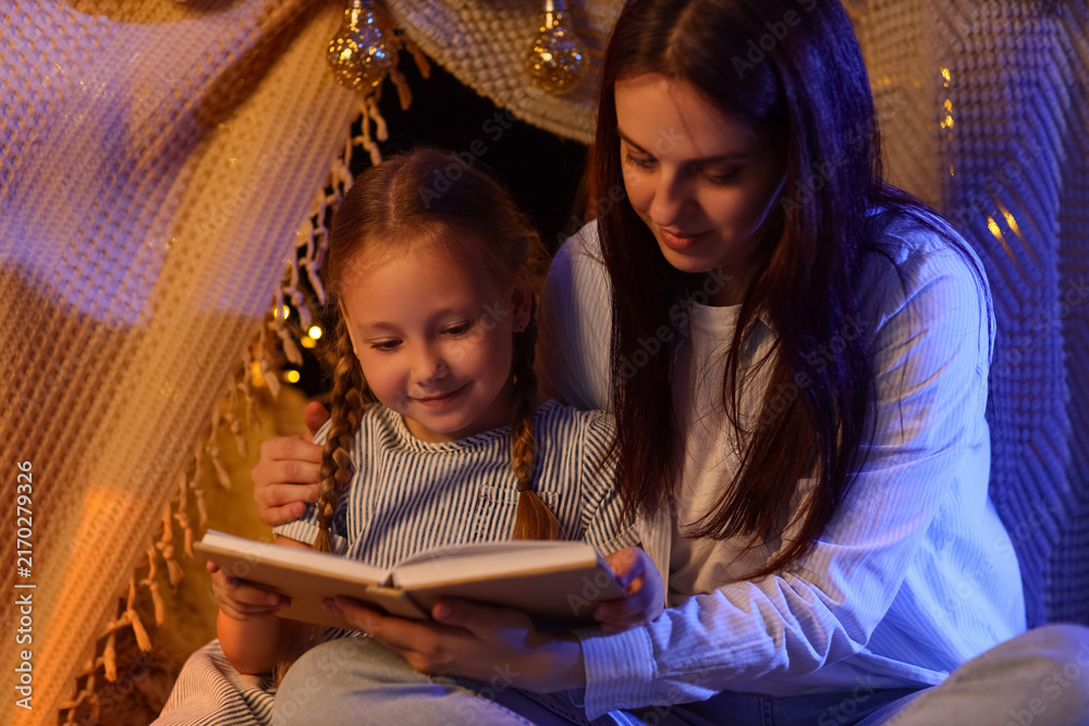 Fototapeta premium Family bonding and childhood development, mother reading a book to her daughter in a cozy blanket fort at night