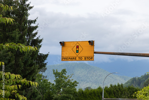 Yellow overhead PREPARE TO STOP warning sign with unlit amber beacons and a signal ahead symbol, set against evergreen forest, misty mountains and a pale overcast sky.
