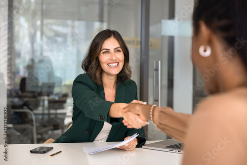 Mexican businesslady, HR manager greeting female African, vacancy applicant with handshake gesture, seated at office desk with resume paper and laptop, starting job interview, feel satisfied. Staffing