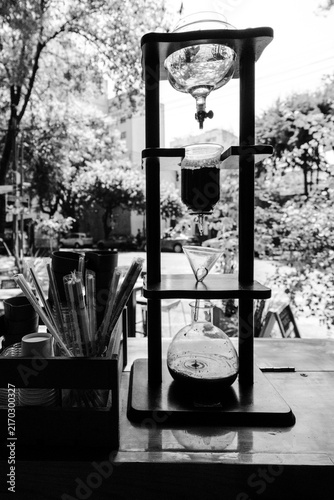 A black and white portrait of a Yama glass cold coffee drip maker on a counter of a coffee shop
