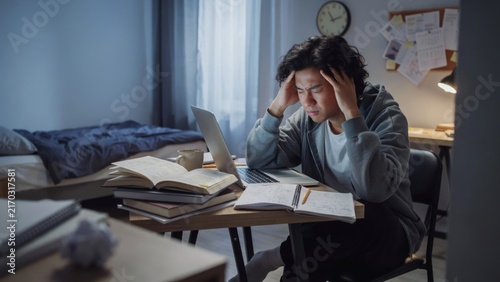 Stressed young asian student studying late at night with laptop and books in a dimly lit room, feeling overwhelmed and tired