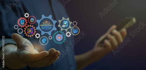 Fotografie Businessman holding smartphone while finger touching glowing gear mechanism with Benchmark central text
