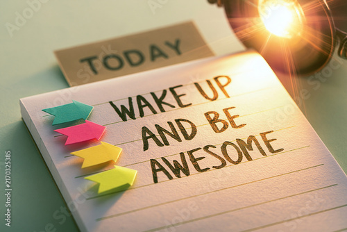 A notepad with handwritten text Wake Up And Be Awesome and colored markers under bright warm light. This represents morning motivation and productivity goals.
