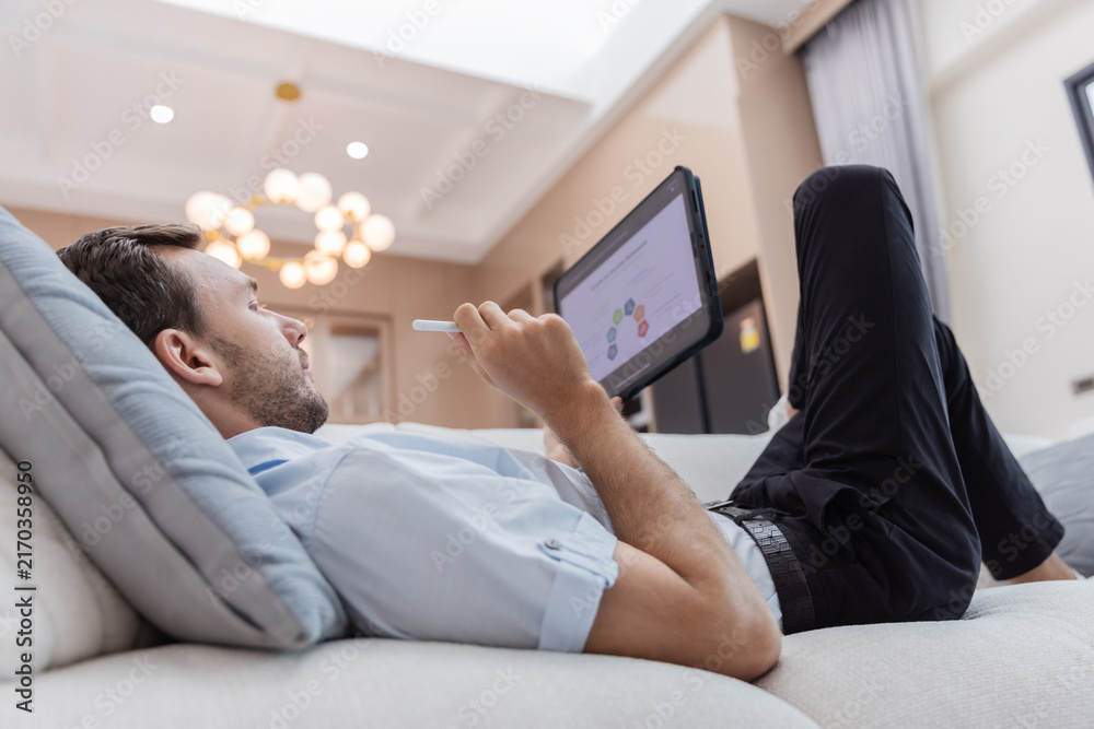 Fototapeta premium Young businessman relaxing on a sofa while reviewing financial charts and business analytics on a digital tablet in a modern living room. 