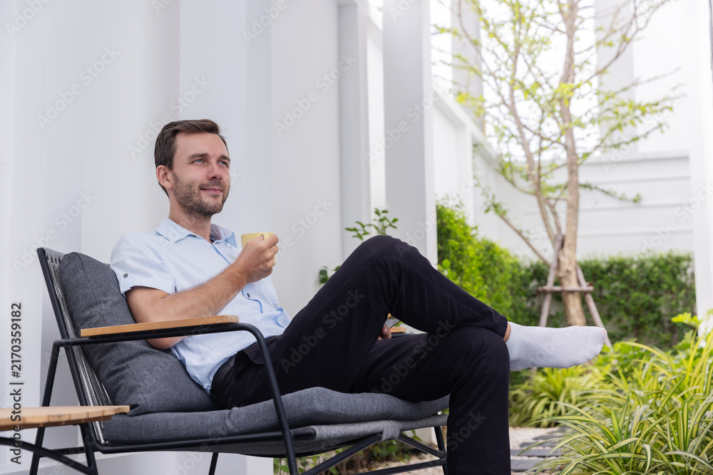 Fototapeta premium Smiling young man enjoying a cup of coffee while relaxing on a comfortable chair in a modern outdoor patio and green garden.