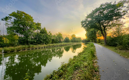 Fotografía golden sunrise over mirrorlike canal surface with towpath and trees, warm light