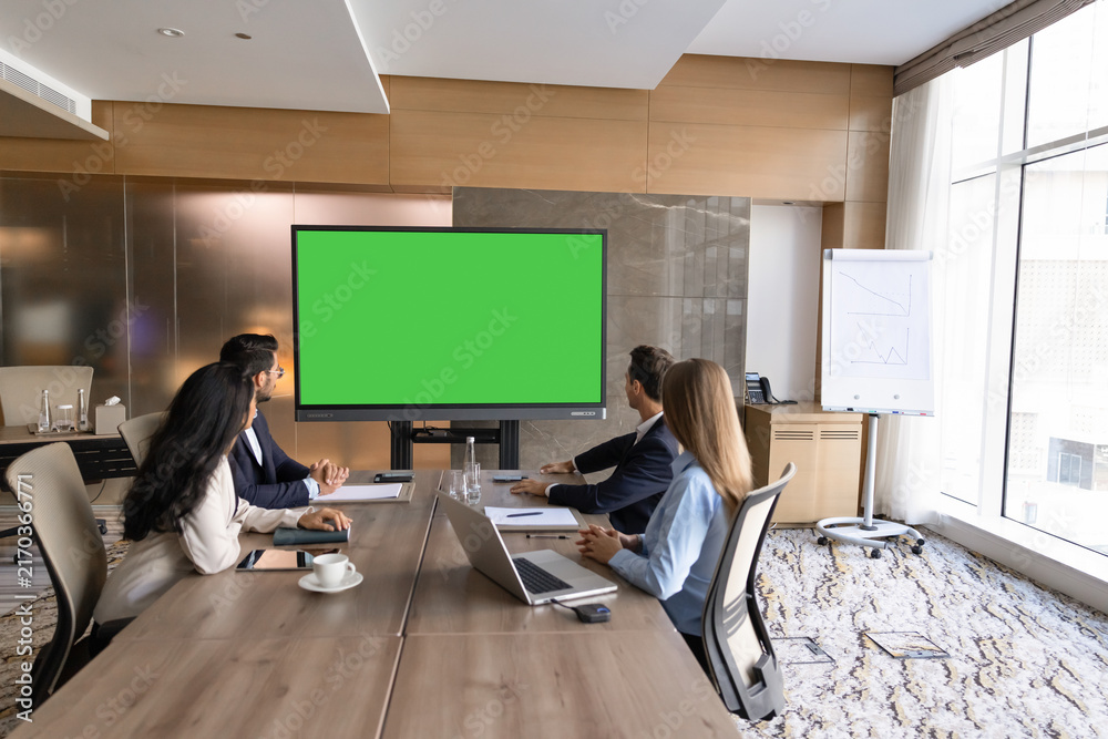 Obraz premium Group of businesspeople sit at conference table, watching presentation, engaging in video call, looking at digital screen displaying green mockup during corporate business meeting in office boardroom