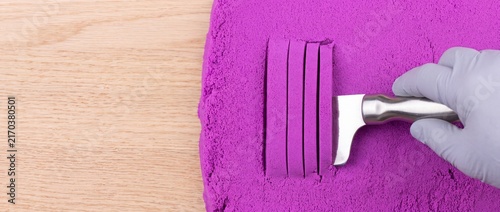 Hand wearing white glove slicing purple kinetic sand into neat strips with a metal spatula on wood.