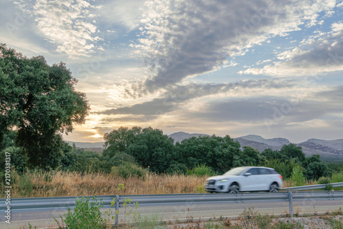 White car speeding on a rural highway during golden hour, motion blur indicating movement, with a scenic backdrop of trees and mountains