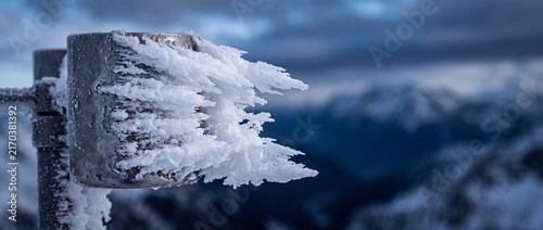 Natural hoarfrost crystals growing on metal pole in alpine mountains against cold winter sky highlights extreme weather freezing.