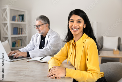 Portrait of South Asian woman patient sits at desk during visit at hospital while male doctor working on laptop, checking medical information, preparing treatment plan. Healthcare consultation concept