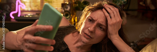 Stressed Caucasian young adult woman looking at smartphone screen with worried expression. Image represents cyberbullying, bad news, or digital fatigue for mental health campaigns