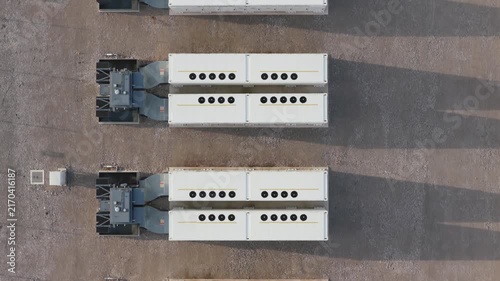 Rows of white industrial battery units occupy a gravel site during the day to store electricity for power distribution and grid management across the expansive outdoor industrial facility