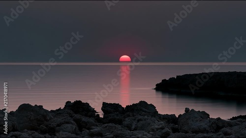 Minimalist Red Sun Setting Over Calm Sea with Dark Rocky Shoreline Silhouette