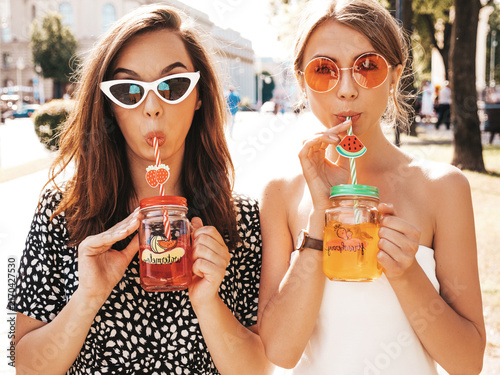 Two young beautiful smiling hipster in trendy summer clothes,dress.Sexy carefree women posing in the street.Positive models having fun in sunglasses.Drinking fresh cocktail smoozy drink