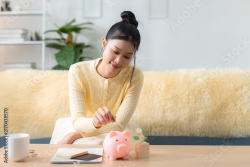 Canvastavla Saving money concept with a young woman putting a coin into a piggy bank while sitting at home, personal finance, savings, budget, investment, planning