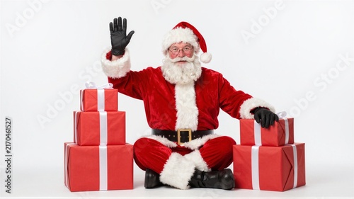 Smiling man in Santa suit waving beside stack of wrapped red gifts on white background