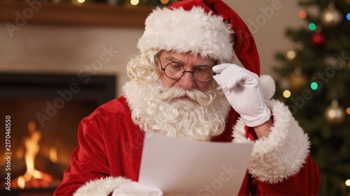 Elderly man in Santa costume reading letter by fireplace with Christmas tree bokeh