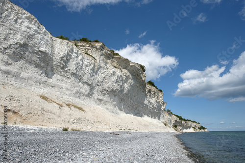 Majestic white chalk cliffs rise steeply from a pebble beach along the turquoise Baltic Sea under a sunny blue sky with clouds on Stevns Klint, Denmark, on a summer day.