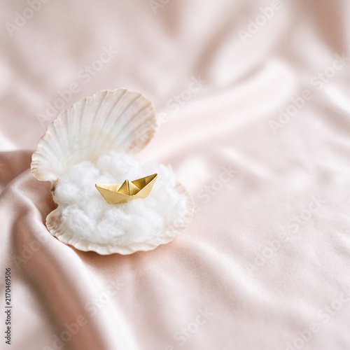 Minimalist surreal still life: golden paper boat on a fluffy cloud inside an open seashell over blush pink satin fabric
