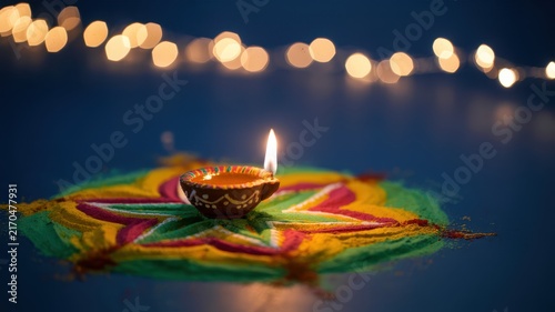 Happy Diwali Single burning clay diya lamp placed on colorful rangoli sand art pattern with bokeh fairy lights on dark blue background