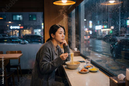 Young woman eating ramen by a rainy window at night for moody restaurant background and city banner design