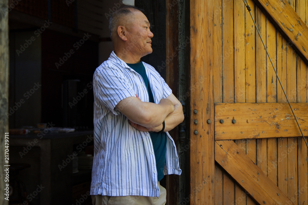 Fototapeta premium Senior Asian man standing in shed doorway by wood door, crossing arms, wearing striped shirt watch