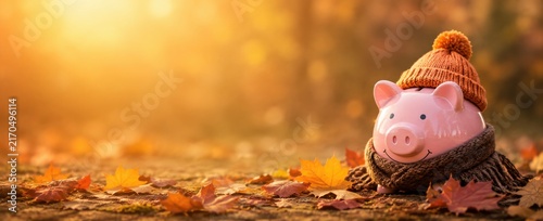 Piggy bank wearing a knitted hat and scarf among fallen autumn leaves, representing saving money, personal finance, budgeting, financial planning and fall season. Advertising banner, copy space