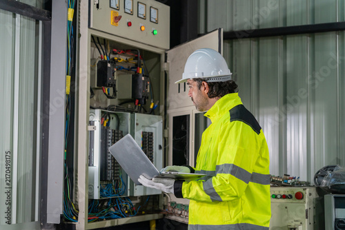 Technician wearing hardhat using laptop computer to inspect electrical control cabinet in factory.