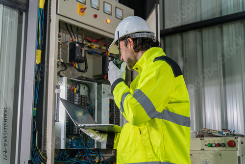 Technician wearing hardhat using laptop computer to inspect electrical control cabinet in factory.