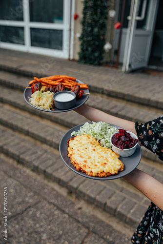 Canvastavla Person carries two plates with cheese-topped breaded cutlets, cabbage salad, bee