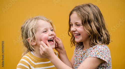 Two happy young girls laughing and playing, one gently squeezing the other's cheeks on a yellow background.