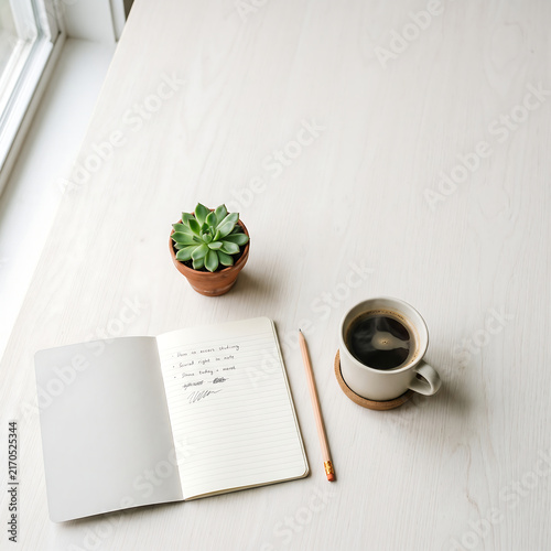 A clean white desk with a to-do list notebook, pencil, coffee cup, and succulent plant bathed in soft morning light.