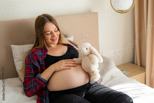 Cuadro en lienzo Smiling expectant mother relaxing on bed with stuffed bunny touching her pregnan