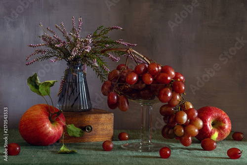 Still life with ripe red apples, sweet grapes and a small bouquet of blooming heather.