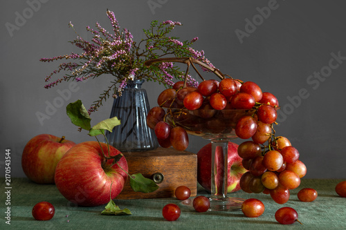 Still life with ripe red apples, sweet grapes and a small bouquet of blooming heather.