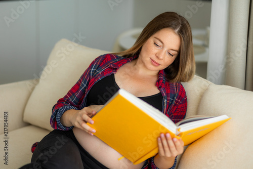 Bild auf Leinwand Pregnant woman reading a yellow book on a comfortable sofa at home, relaxed and