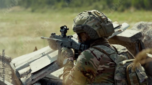 Close-up of armed soldier in full camouflage tactical gear and helmet firing assault rifle from concrete trench position in open field. Military warfare concept for defense, conflict and editorial use