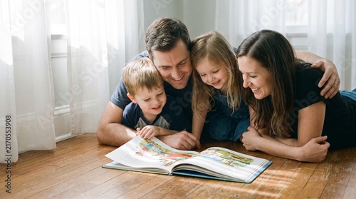 A happy family lying on the wooden floor and reading a book together in the living room.