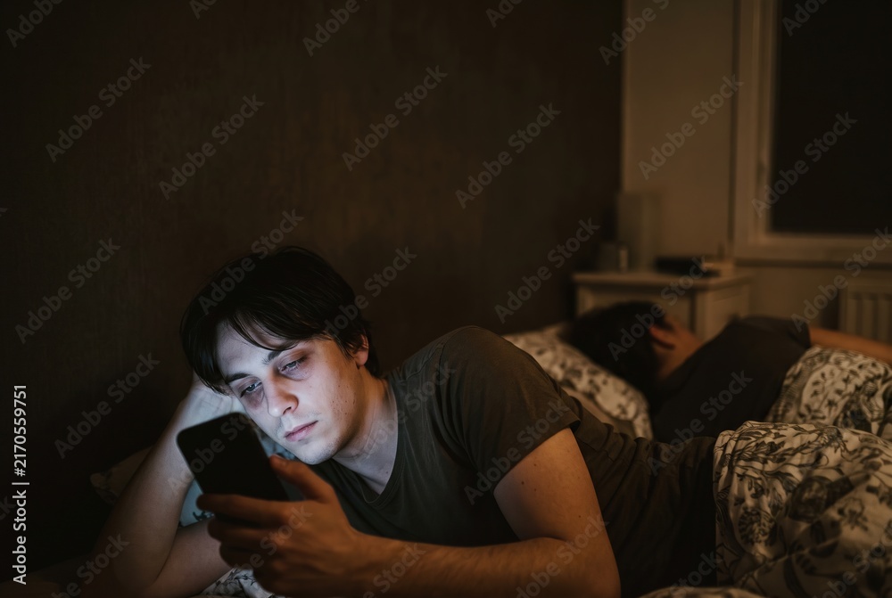 Obraz premium A man is lying in bed at night. He holds his phone and stares at the screen. Another person is lying beside him in the same bed, not looking at the phone