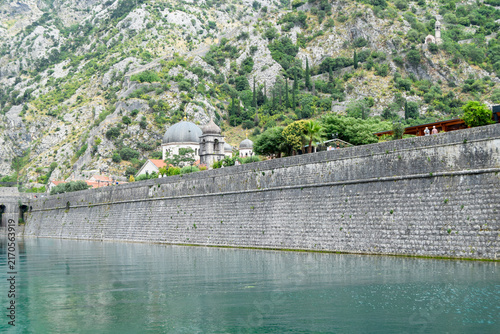 Ancient stone fortress wall and round bastion tower standing by the water. Historical fortification architecture with steep mountain backdrop and coastal heritage detail.