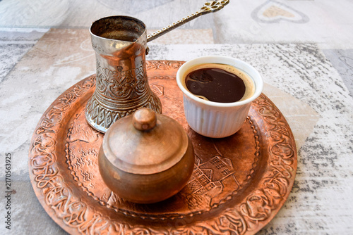 Traditional Turkish coffee served in a white cup on an engraved copper tray alongside an authentic cezve pot and copper sugar bowl. Oriental coffee culture and authentic copper craft detail.