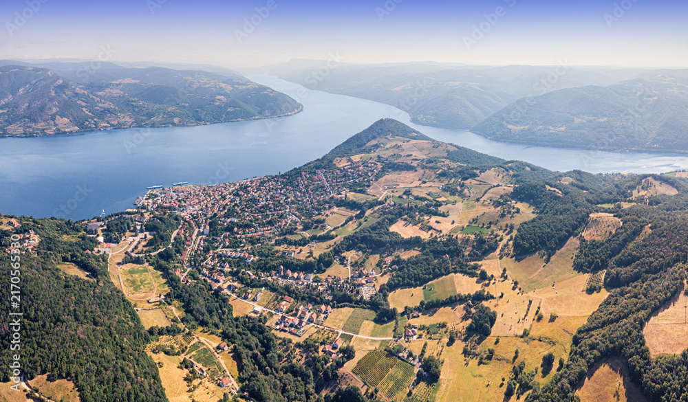 Fototapeta premium Bright summer aerial panorama of a riverside town nestled among rolling green hills along a wide Danube river winding through forested valleys. Travel concept.