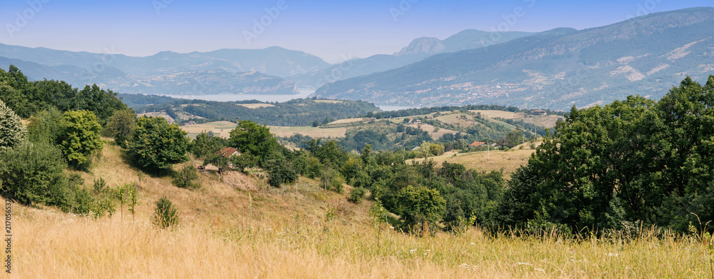 Naklejka premium Panoramic hillside view sweeping across layered green valleys, golden meadow, scattered farmhouses, and a distant Danube framed by misty mountains. Travel concept.