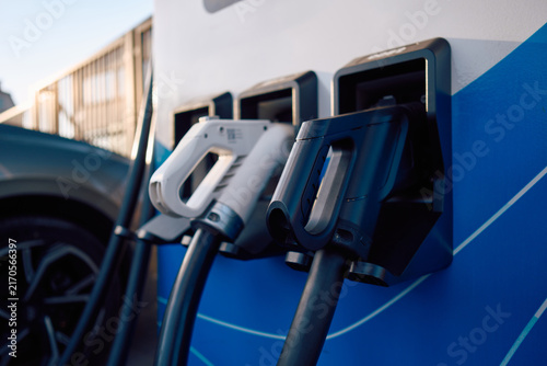 Tight shallow-depth view showing multiple electric vehicle charging nozzles docked in holders on blue public charging station, black and white ergonomic handles, thick cables, urban energy