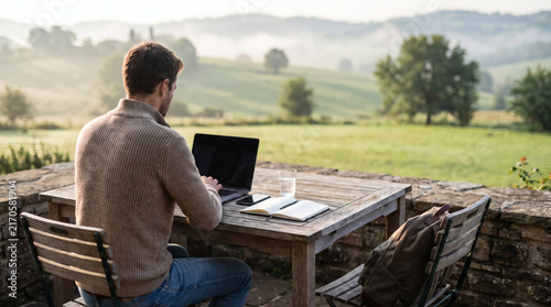 young man working remotely on laptop at outdoor terrace table with scenic mountain landscape and rural view