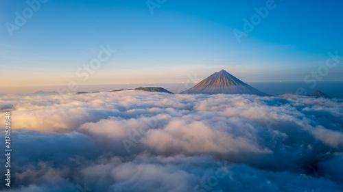 Aerial view of Mount Inerie rising majestically above a dense sea of clouds under a clear blue sky in the morning light near Bajawa, Nusa Tenggara Timur, Indonesia.
