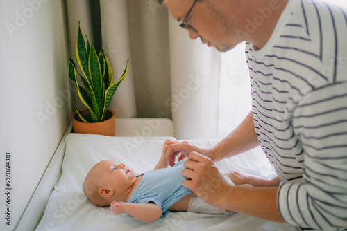 Bild auf Leinwand Young father carefully dressing his infant son on a white changing table, puttin