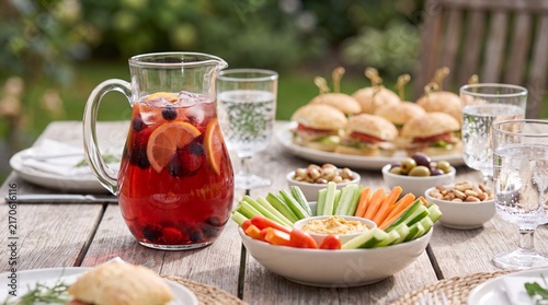 Refreshing pitcher of fruit-infused beverage with sliced oranges and berries on a wooden table, surrounded by plates of sandwiches and fresh vegetables